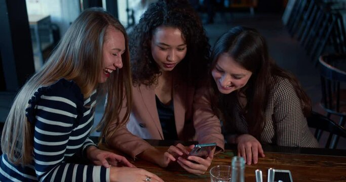 Social media concept. Three young beautiful happy multiethnic female friends using smartphone together at coffee shop.