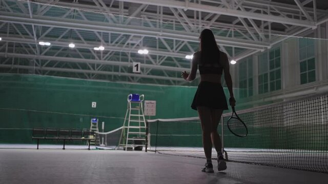 young female tennis player on pro competiton, walking on tennis court, holding tennis racquet and throwing ball, rear view