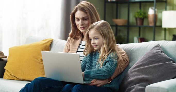 Happy mother and cute little daughter doing interesting tasks together using laptop sitting at home on couch.
