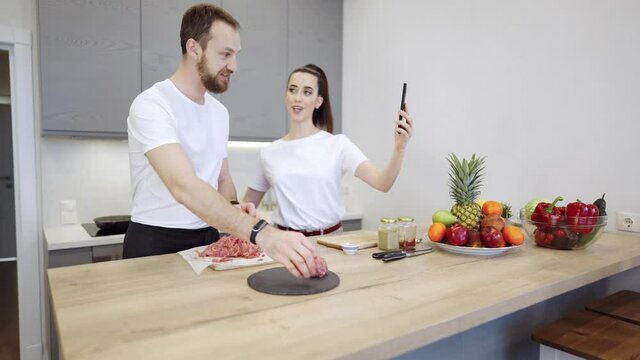 Young cheerful couple cooking food together. They are preparing meat burgers at home in the kitchen. girl has a video conference or chat with a smartphone, saying hi