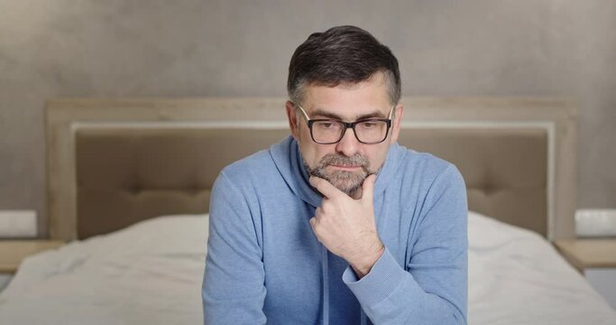 Serious look at camera senior man frustrated upset by bad news emotionless apathy depression sitting at home on bed in bedroom