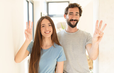 young couple smiling and looking friendly, showing number three or third with hand forward,...