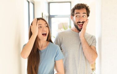 young couple feeling happy, excited and surprised, looking to the side with both hands on face