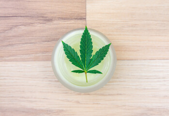 Flat lay of CBD lotion in glass container with Cannabis leaf on wooden backdrop