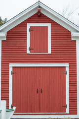 An old two storey red barn with large double red doors, white trim, and a small closed wood loft window. The sky in the background is grey. There's a wooden boat on the ground in front of the doors. 