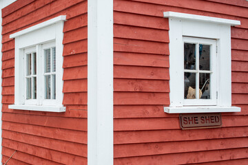 Fototapeta na wymiar A small closed glass window with wide white trim. The exterior wall of the building is a bright red narrow wood clapboard. There's a sign with the words she shed under the window ledge.
