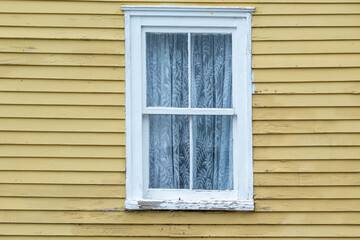 The exterior of a vintage yellow coloured wall with narrow wood cape cod clapboard siding. In the center of the house, there's a closed double hung window with a white lace curtain. 