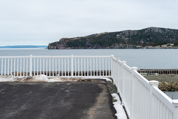 Obraz premium A long white metal fence and corner join with post caps along the edge of a paved parking lot overlooking the blue ocean. There's a tree covered island in the background under white cloudy skies. 