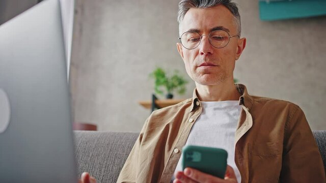 Two factor authentication.Mature man in eyeglasses entering security password from smartphone to website on laptop