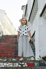 Naklejka premium A stylish model, a six-seven-year-old girl with blond hair, stands on a bright stone staircase in a city street.