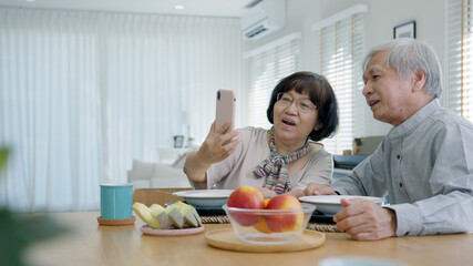 Young senior asia citizen couple hold mobile phone selfie videocall online to family at home apartment morning on dining table in older people with digital technology virtual talk conference concept.