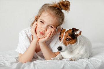 Beautiful funny kid girl lies in bed with a dog in a white bedroom.