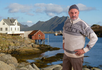 An elderly Norwegian with a beard and a typical sweater is standing on the bank near Tromso. In the background is a fishing boat and mountains. On the left is a Norwegian wooden house built on stilts.