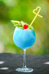 Vertical picture of blue cocktail with apple, cherry and yellow straw in wine glass on background of green bokeh. Bartender skills concept 
