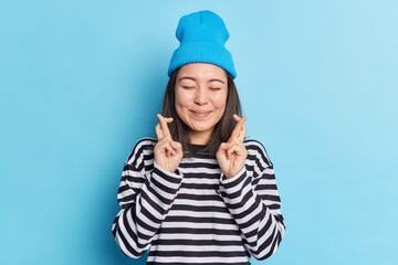 Happy pleased Asian teenage girl crosses fingers anticipates for results keeps eyes shut believes dreams come true wears casual striped jumper and hat isolated over blue background. Good luck