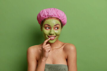 Happy dreamy young woman stands wrapped in bath towel shows bare shoulders smiles toothily cares...