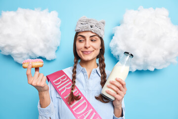 Pleased European woman with two pigtails looks at tasty glazed doughnut going to eat it with fresh milk likes sweet delicious desserts wears domestic clothes isolated over blue background. Temptation