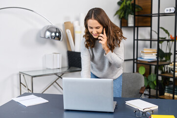 Busy woman talking with a customers on the phone and types on the keyboard, standing near office desk, multitasking female office employee