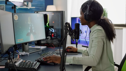 Competitive black gamer with headphones using stream chat on RGB powerful computer playing online shooter game late at night. Virtual online streaming cyber performing in gaming tournament