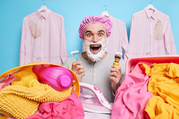 Positive surprised bearded man applies foam gel for shaving holds razor and brush does daily morning routines irons linen in laundry room wears hat for taking shower isolated over blue background