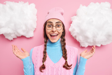 Positive millennial girl raises palms reacts on something awesome smiles broadly wears round eyeglasses hat and vest hears excellent news express good emotions isolated over pink background.