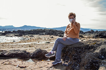 senior woman with mobile phone on the beach and wearing disposable mask. Security measures during the coronavirus outbreak