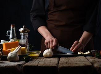 Chef slices garlic for preparing a dish of meat and salad. On the background of ingredients, cheeses, olive oil and spices. Recipes and food preparation, illustration for the menu book