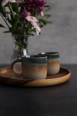 Two elegant rustic coffee cups on a wooden tray next to a blurred bouquet of dahlias.