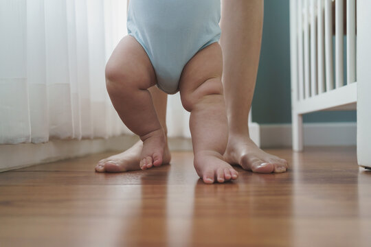 Asian Mother Holding Her Little Baby Hands Learning To Walk On Wooden Floor At Home. Cute Toddler Enjoying The First Steps With Mom.