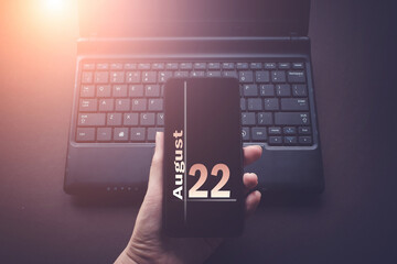 August 22nd. Day 22 of month, Calendar date. Hand Holding Mobile Phone on Laptop Computer dark background with sunshine. Summer month, day of the year concept.