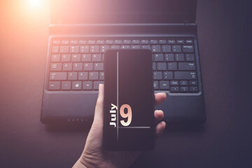 July 9th. Day 9 of month, Calendar date. Hand Holding Mobile Phone on Laptop Computer dark...