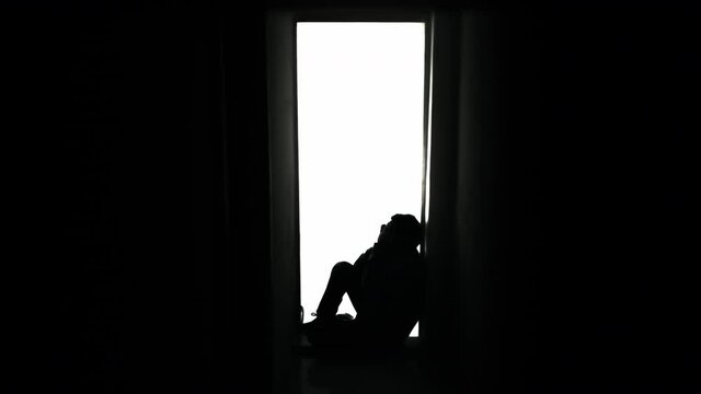 A sad man, sitting on a doorway in his house, crying alone. Silhouetted shot.

