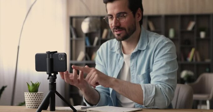 Focus on modern gadget smartphone standing on tripod stabilizer on table, recording skilled young businessman teacher tutor on web camera. Confident male blogger mentor filming educational video.