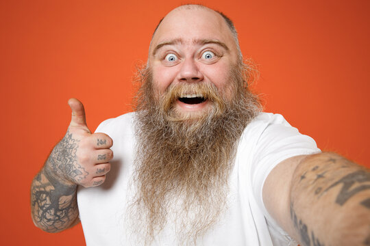 Close up fat pudge obese chubby overweight tattooed blue-eyed bearded man in white t-shirt do selfie shot on mobile phone show thumb up like gesture isolated on red orange background studio portrait.