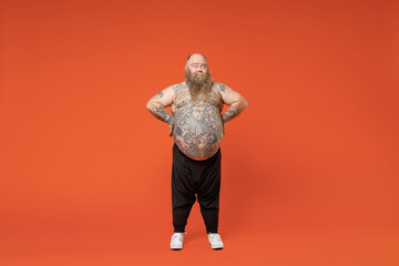 Full length fat fun cool caucasian pudge obese chubby overweight tattooed blue-eyed bearded man 30s has big belly in black pants naked torso stand akimbo isolated on orange background studio portrait.