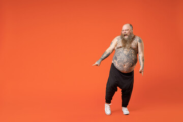 Full length fat fun overjoyed joyful pudge obese chubby overweight tattooed bearded big belly man 30s wear black pants naked torso dancing fooling around isolated on orange background studio portrait.