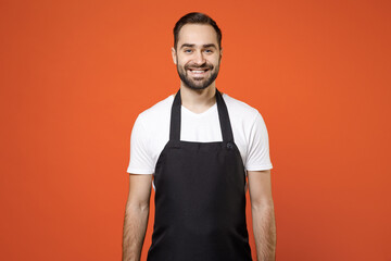 Young fun man 20s barista bartender barman employee in black apron white t-shirt work in coffee shop looking camera isolated on orange color background studio portrait. Small business startup concept.
