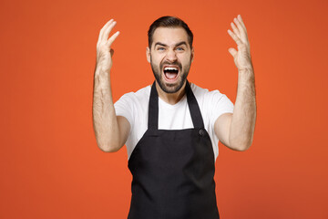 Young indignant angry stressed man barista bartender barman employee in black apron white t-shirt work in coffee spread hands scream shout isolated on orange background studio Small business startup.