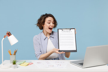 Young surprised successful employee business woman in casual shirt sit work at white office desk with pc laptop hold clipboard with papers document isolated on pastel blue background studio portrait.