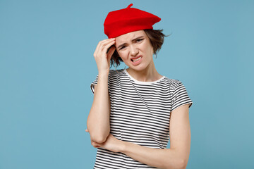 Young sad troubled pensive thoughtful puzzled confused woman 20s, short hairdo in french beret red hat striped t-shirt look aside prop up forehead isolated on pastel blue background studio portrait