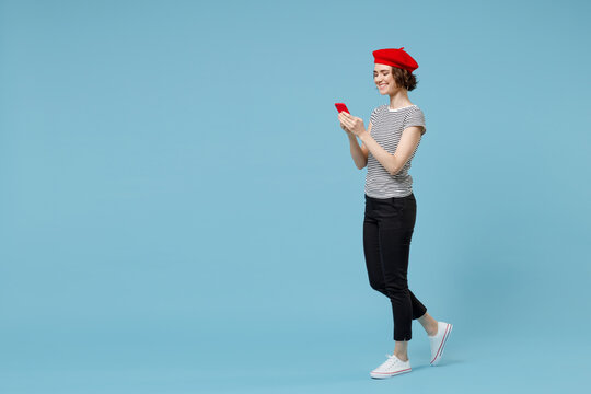 Side Profile View Young Fun Smiling Happy Woman 20s With Short Hairdo In French Beret Red Hat Striped T-shirt Using Mobile Cell Phone Walking Going Isolated On Pastel Blue Background Studio Portrait