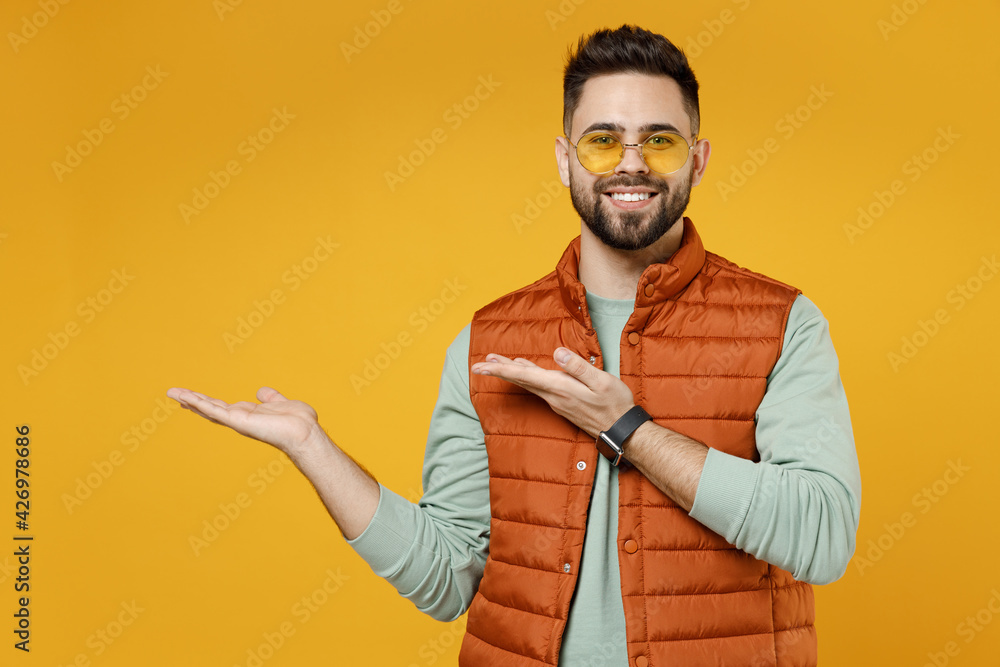 Wall mural Young smiling happy fun man 20s in orange vest mint sweatshirt glasses point arms hands aside on copy space area mock up workspace isolated on yellow background studio portrait. Advertisement concept.