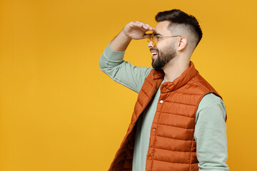 Side profile view expectant young happy fun man 20s years old wearing orange vest mint sweatshirt glasses hold hand at forehead look far away distance isolated on yellow background studio portrait