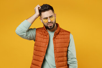 Young puzzled pensive troubled thoughtful man 20s wearing orange vest mint sweatshirt glasses looking aside sctratch hold head isolated on yellow color background studio portrait. Lifestyle concept.