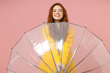 Redhead young woman in yellow waterproof raincoat hood outerwear hiding transparent umbrella isolated on pastel pink color background studio portrait Outdoor lifestyle wet fall weather season concept