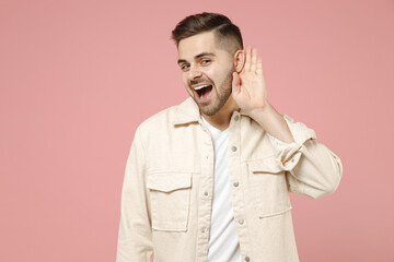 Young curious nosy caucasian fun man 20s wearing jacket white t-shirt try to hearing you overhear listening intently isolated on pastel pink color background studio portrait. People lifestyle concept.