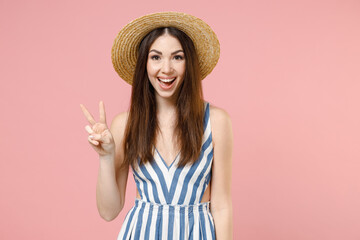Young excited positive cheerful caucasian woman 20s in summer clothes striped dress straw hat show victory v-sign gesture isolated on pastel pink background studio portrait. People lifestyle concept.