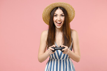 Young happy overjoyed excited caucasian smiling woman 20s wearing summer clothes striped dress straw hat play pc game with joystick console isolated on pastel pink background studio postrait portrait.