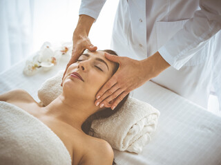 Obraz na płótnie Canvas Beautiful brunette woman enjoying facial massage with closed eyes comfortable and blissful. Relaxing treatment in medicine and spa center concepts