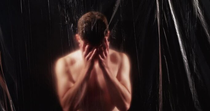 Depressed man. Defocused portrait. Embarrassment regret sorrow. Sad ashamed shirtless guy covering face with hands behind wrinkled texture polyethylene film isolated on black background out of focus.